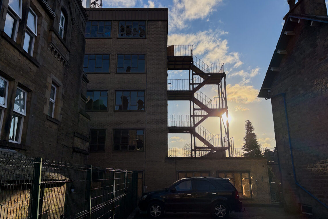 A black SUV is parked beside a fenced building. The sun is setting behind a brick building with metal staircases. Shadows and reflections are visible on the windows, and the sky is partly cloudy with yellow and blue hues.