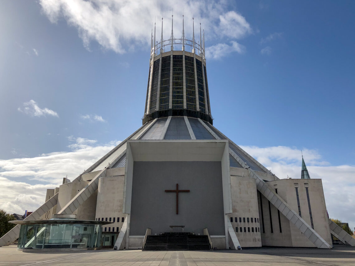 A modern cathedral with a circular tower topped by spires, wide concrete steps leading to a large entrance with a cross above, and a glass-walled structure to the left. The sky is mostly clear with patches of cloud.