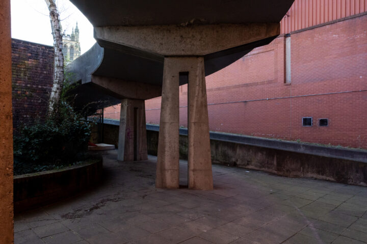 Concrete walkway with angular pillars underneath, casting shadows on paved ground. Brick walls on both sides; a round planter with a tree on the left. In the background, a church tower is partially visible above the wall. The scene appears empty and quiet.