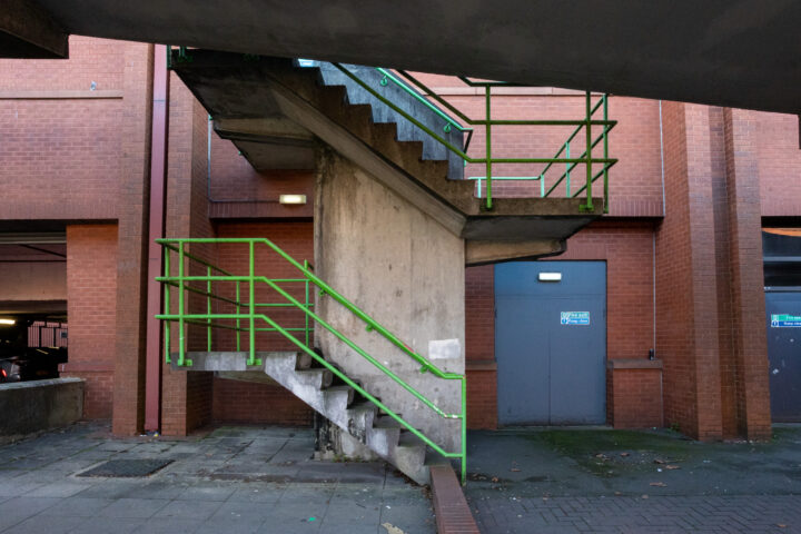 Concrete steps with green railings lead up to a landing outside a red brick building. A grey door with a warning sign and light is on the right. The area is empty, with some moss and fallen leaves on the ground. Shadows fall under the overhang.