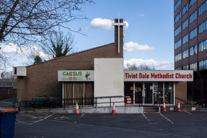 A brick building houses Cactus Café and Tiviot Dale Methodist Church. A cross tops a chimney. Orange cones line a ramp and car park. A tall office block stands to the right. Leafless tree branches frame the left side.