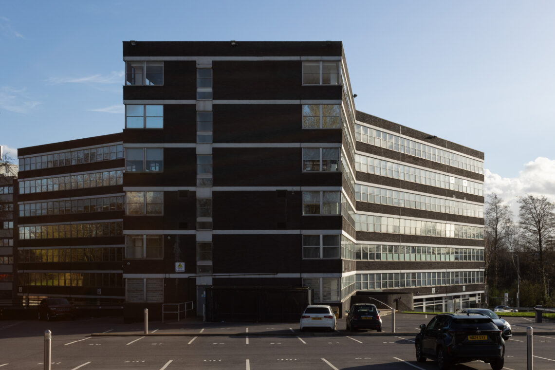 A large, dark brick office building with horizontal white stripes and rows of windows stands in sunlight. Several cars are parked in an almost empty car park in front of the building. The sky is clear with a few clouds, and some trees are visible.