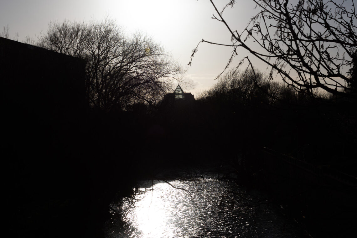 Bare winter trees line a river, reflecting bright sunlight. The image is mostly in shadow, with a silhouetted building featuring a triangular roof in the background against a pale sky.