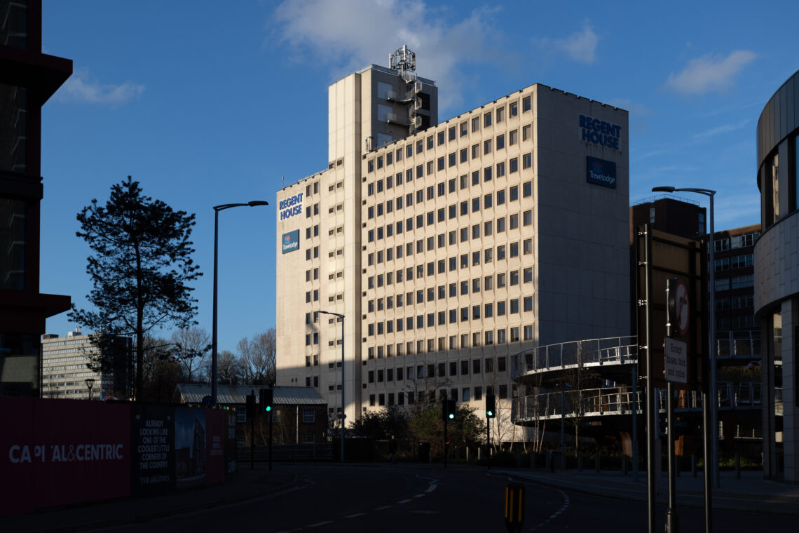 A tall, cream-coloured office building labelled Regent House stands in sunlight, casting long shadows. The street in front is mostly empty, with some trees, streetlights, and signage visible. The sky is clear and blue.