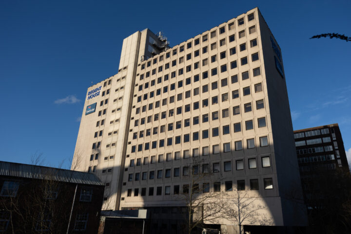 A tall, rectangular beige office building with many square windows is shown under a clear blue sky. The sign on the upper left reads Ingeus House. Shadows from trees and nearby buildings partially cover the lower part of the structure.