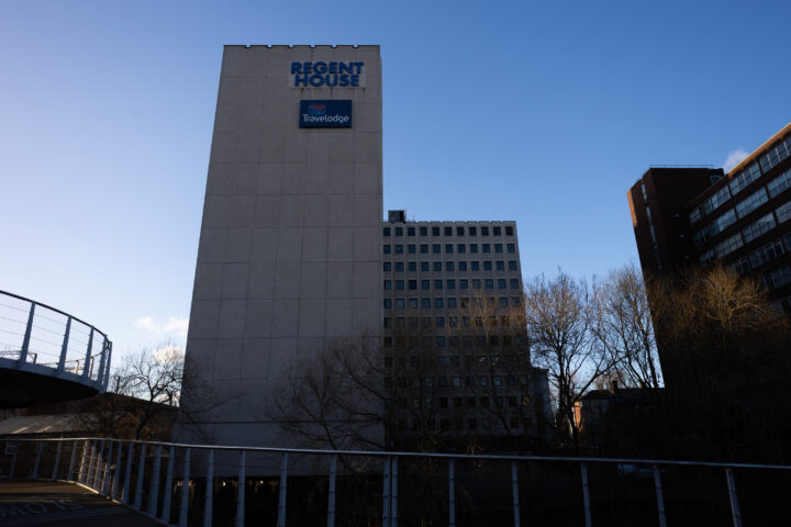 A tall, white building labelled Regent House with a Travelodge sign stands against a clear blue sky. Bare trees and two other buildings surround it. A curved walkway with metal railings appears on the left side of the image in the foreground.