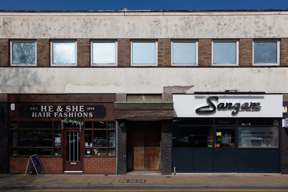 A two-storey building with three shops: He & She Hair Fashions (left, brown sign), an empty entrance (centre), and Sangar (right, white sign). The upper level has six windows; the facade is dirty and weathered. The pavement is visible.