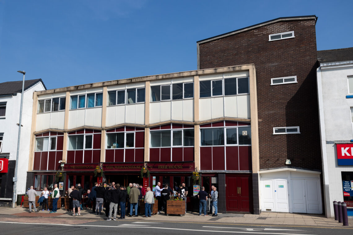 A group of people gathers outside a red and cream three-storey pub on a sunny day. The building has large windows and a sign reading Free House. Some people stand near a wooden barrel. Nearby buildings and a road are visible.