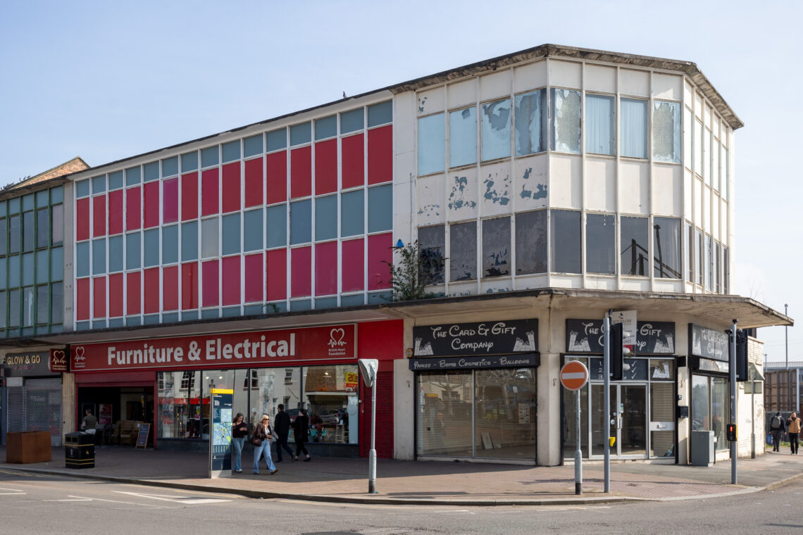 A corner building with faded red and white panels, peeling upper windows, and shops below—a Furniture & Electrical shop and a Card & Gift shop. Several people walk by on the pavement under sunny skies. A no entry road sign is nearby.