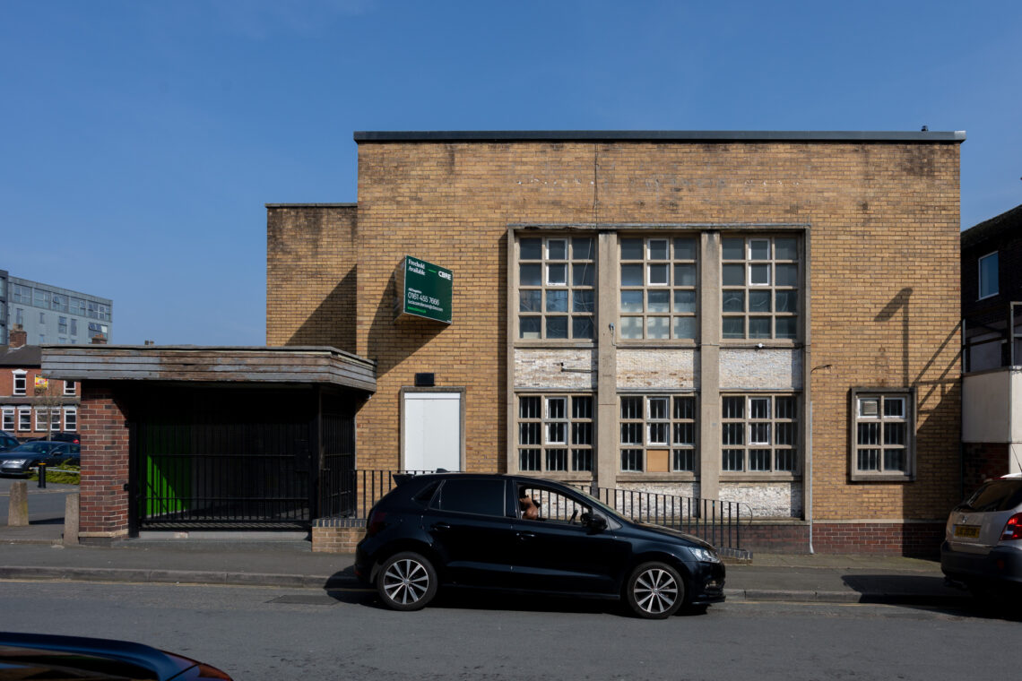 A small brick building with tall, old windows stands on a sunny street. A black car with a person driving passes by in front. There’s a wooden bus shelter to the left, and other parked cars to the right. The sky is clear and blue.