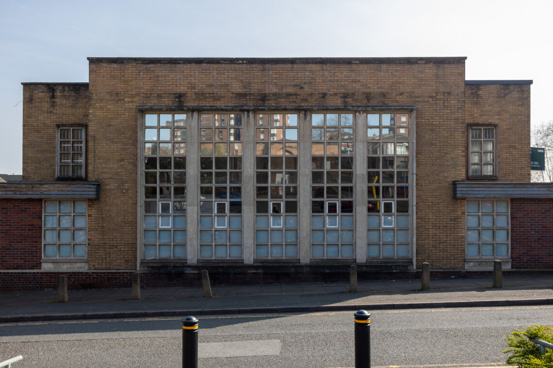 A brown brick building with large rectangular glass windows arranged in a grid, flat roof, and short wings on each side. Three black and yellow bollards and a small step are at the roadside in front. Reflections of other buildings are visible in the windows.