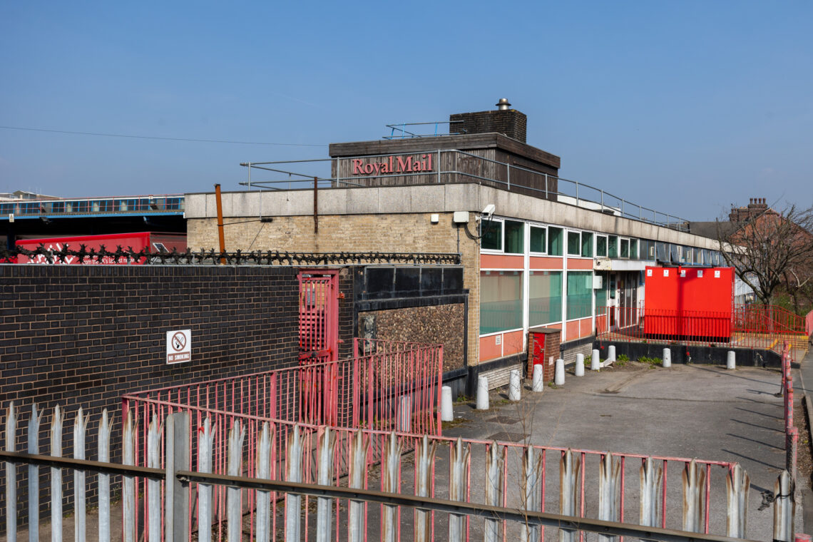 A fenced Royal Mail building with barred windows, red doors, and a No Smoking sign. The yard is empty, bordered by red-and-white railings and bollards. The sky is clear and blue.