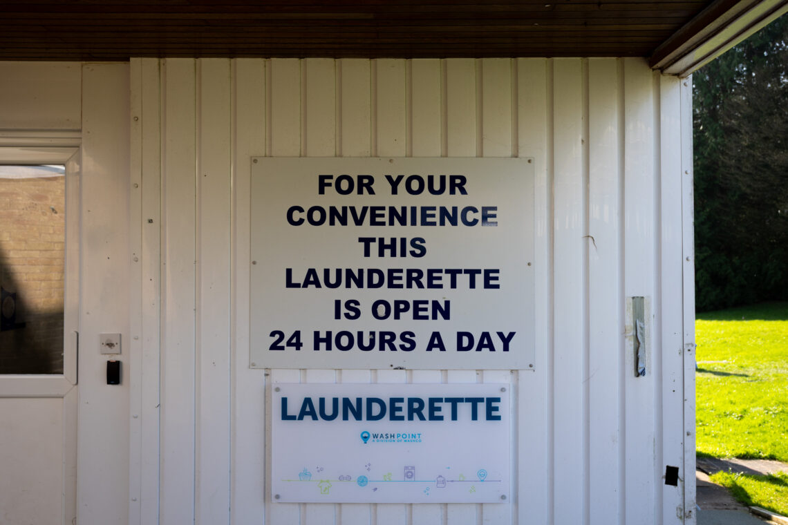 A white building with two signs; the top says, For your convenience this launderette is open 24 hours a day in bold black letters, and the lower sign reads, Launderette Wash Point with blue icons, next to a grass lawn and trees.