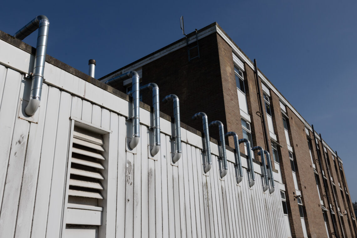 A row of shiny metal pipes lines the top of a white wooden fence in front of a brick building with multiple windows, under a clear blue sky.