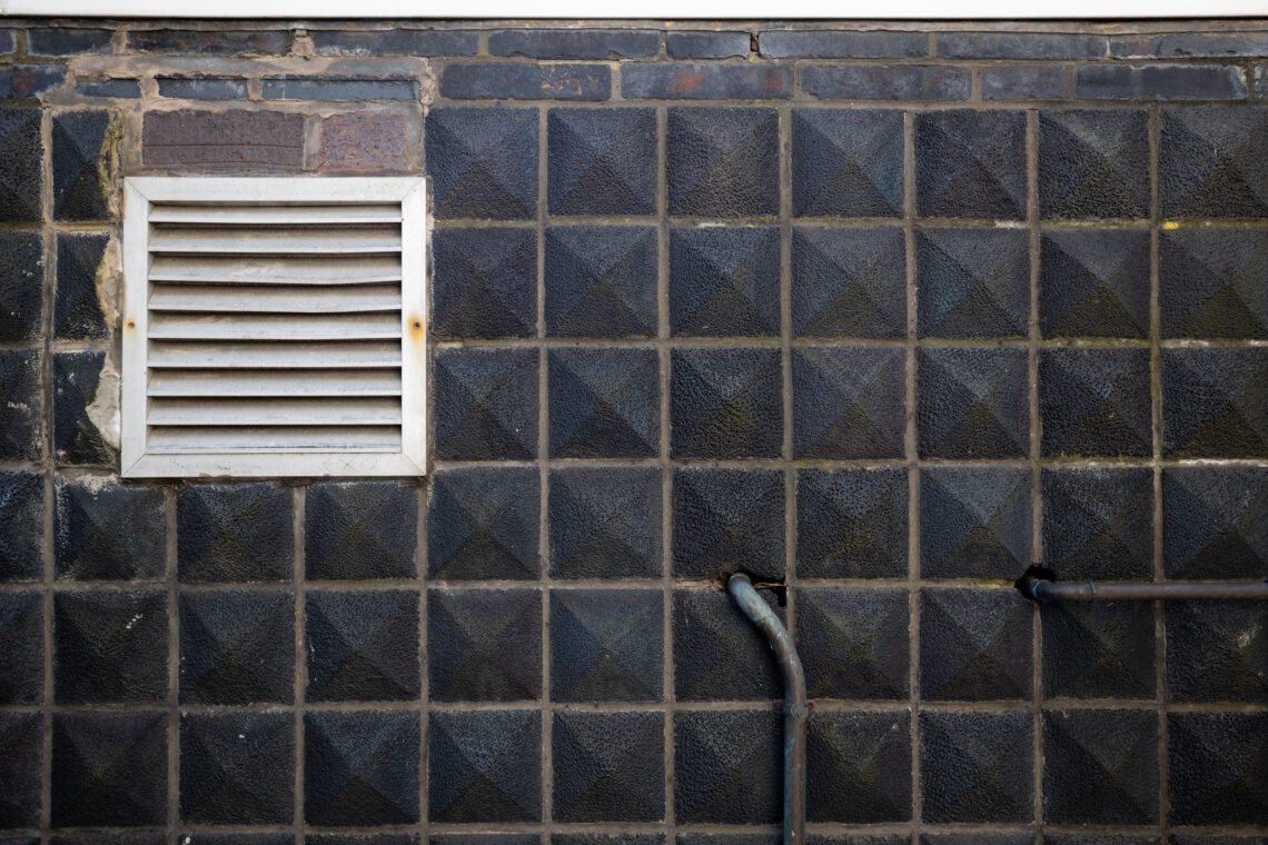 A textured black wall with a grid of raised diamond patterns has a white metal vent in the upper left and two protruding metal pipes near the lower right. Bricks and a white trim are visible at the top edge.