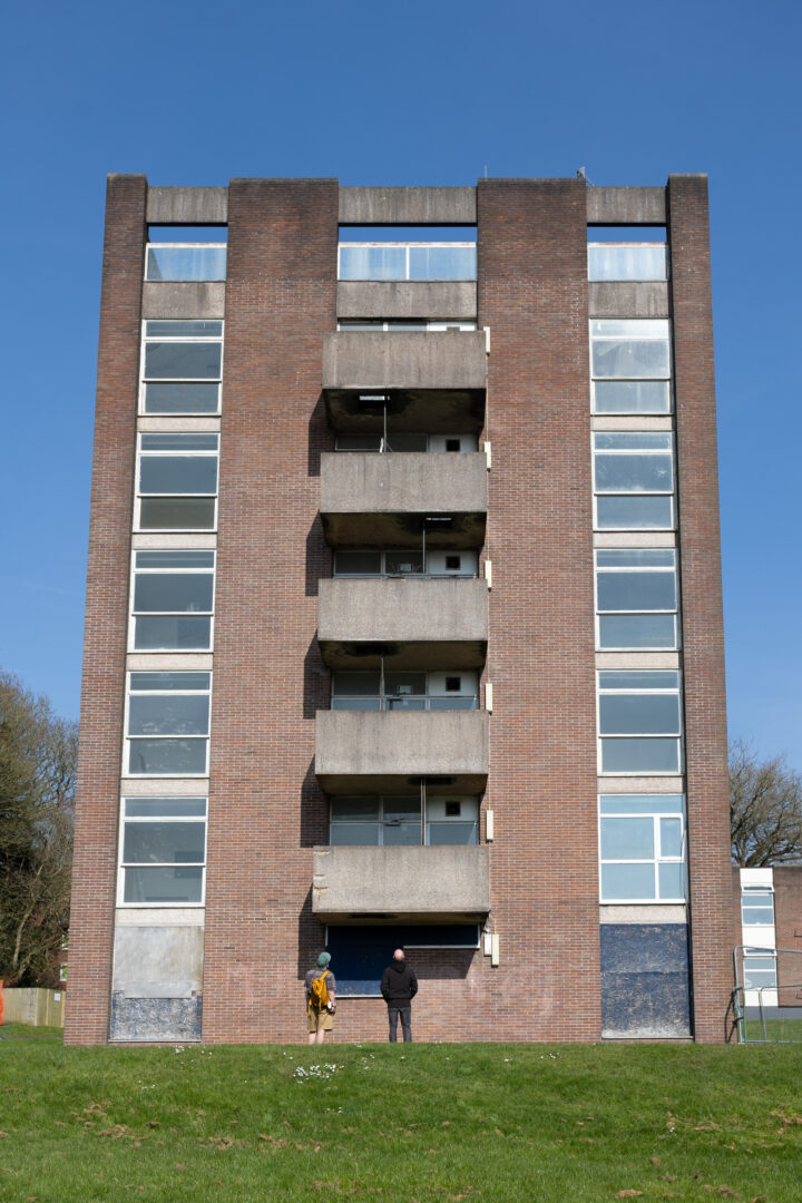 A tall, rectangular brick block of flats with balconies and windows stands on green grass. Two people, one in yellow and one in black, face the building's entrance. The sky is clear and blue.