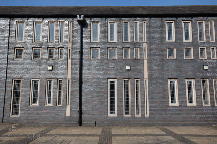 A brick building with many tall, narrow windows arranged in rows, each with white frames. A black drainpipe runs vertically between the windows. The ground is paved with large stone slabs. The sky is clear and blue.