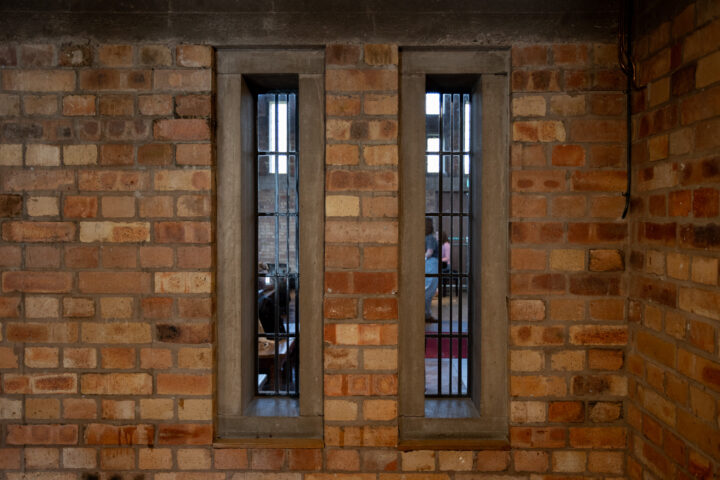 Two tall, narrow windows with metal bars are set in a brick wall. Through the windows, blurred figures and a table are visible, suggesting people seated in a room beyond. The bricks vary in colour, giving the wall a textured, rustic appearance.