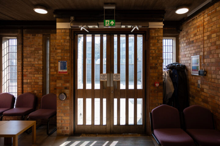 A bright room with purple chairs, a small table, and a coat stand faces large wooden double doors with barred windows. Sunlight streams in, and emergency exit signs are above the doors. Brick walls wrap around the cosy waiting area.
