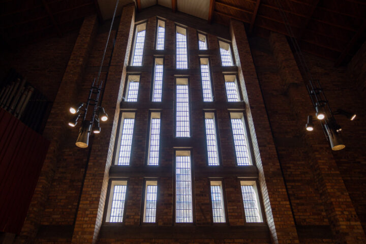 Tall, multi-panelled window with geometric vertical sections set in a brick wall inside a building with high ceilings. Modern hanging light fittings are visible on both sides. Sunlight streams through the glass, illuminating the interior.