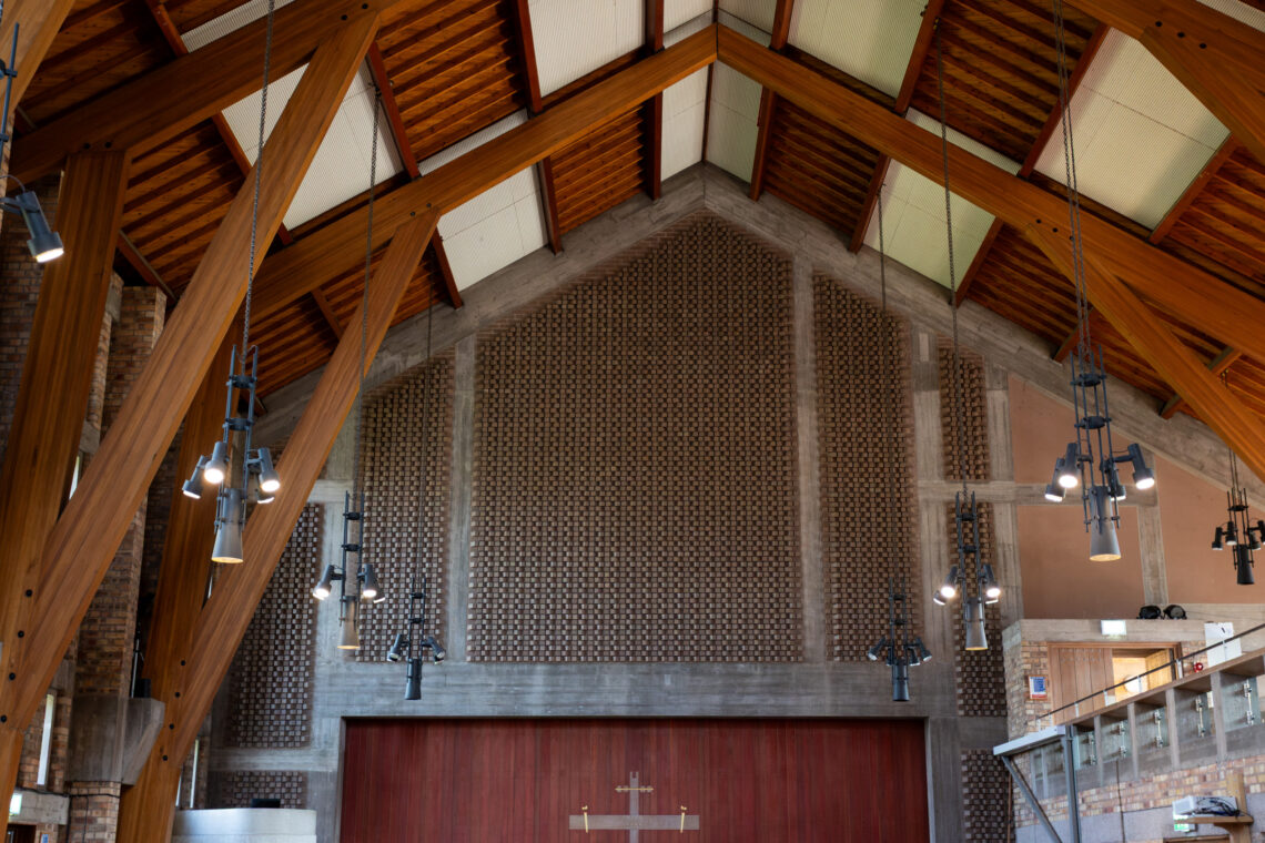 Interior of a modern church with a high wooden ceiling, exposed beams, and pendant lights. The back wall features a cross, textured panels, and a large wooden door. Glass railing and people are visible on the right. Warm natural light fills the space.