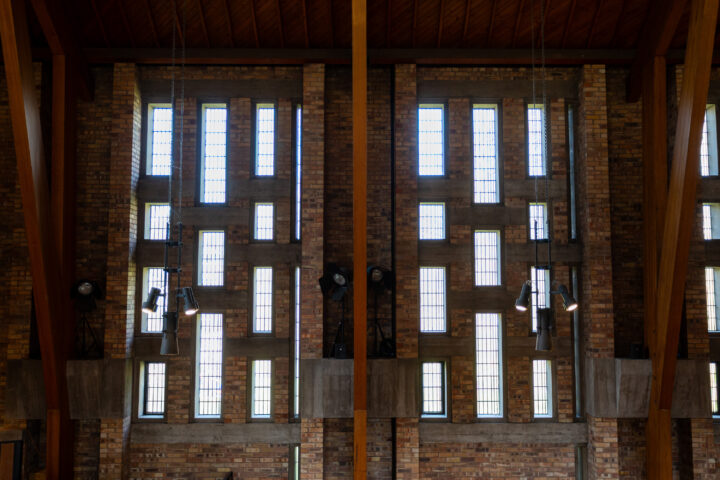 Tall brick wall with three large vertical sections of rectangular glass windows. Wooden beams divide the sections. Modern stage lights are mounted near the windows. Light streams in, illuminating the interior. Ceiling is angled and has a wooden finish.
