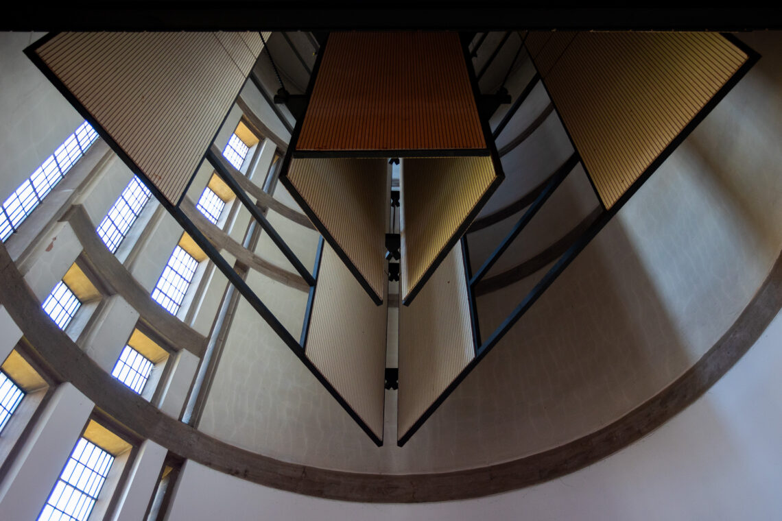 View looking up at a modern architectural ceiling with geometric acoustic panels suspended in rows, surrounded by curved concrete beams and tall windows with rectangular glass panes letting in natural light.