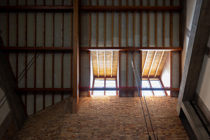 Interior view looking up at a ceiling with exposed wooden beams, two large windows letting in daylight, a brick wall below, red pipe, and concrete pillars to the right; the setting suggests an industrial or loft-style space.