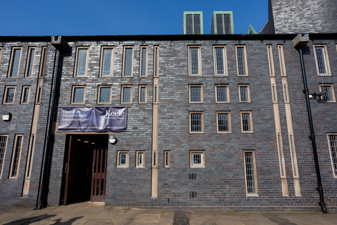 A modern brick building with tall, narrow windows. A purple banner above the door reads, Open for all. Free to enter. Everyone welcome. Keele Chapel. The entrance is open, and the sky is clear and blue.