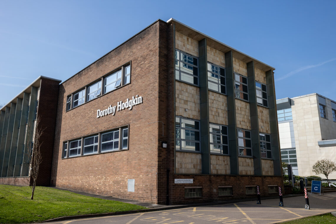 A modern, rectangular brick building with large windows, labelled Dorothy Hodgkin. It sits under a clear blue sky. The surrounding area has grass, a pavement, a small tree, bollards, and sections of adjacent buildings visible.