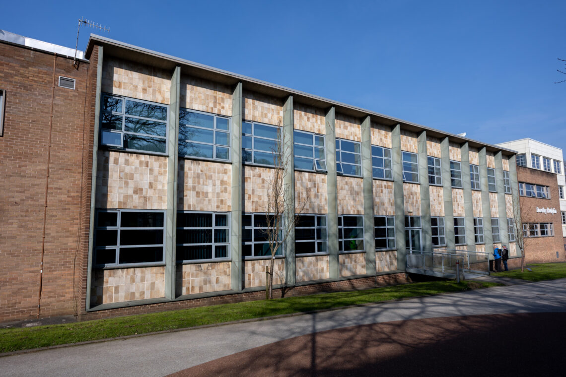 A two-storey building with a brick façade and large rectangular windows, divided by vertical concrete pillars. A small ramp leads to an entrance on the right. Two people stand near the door. The sky is clear and blue, with green grass in front.