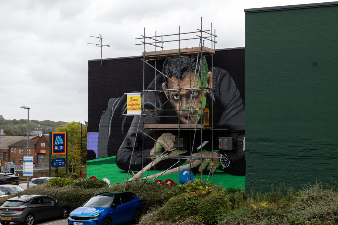 A mural of a man playing snooker is painted on a building’s side. Scaffolding partially covers his face and arm. An Aldi store sign, cars, greenery, and a cloudy sky are visible in the scene.