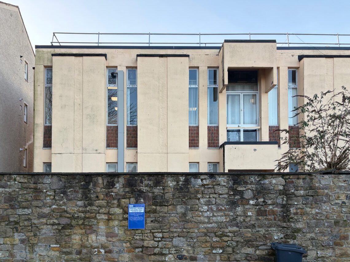 A beige, two-storey building with vertical panels and narrow windows stands behind a stone wall. A blue sign is attached to the wall, and a rubbish bin is visible in front. Some plants grow to the right. The sky is clear and blue.