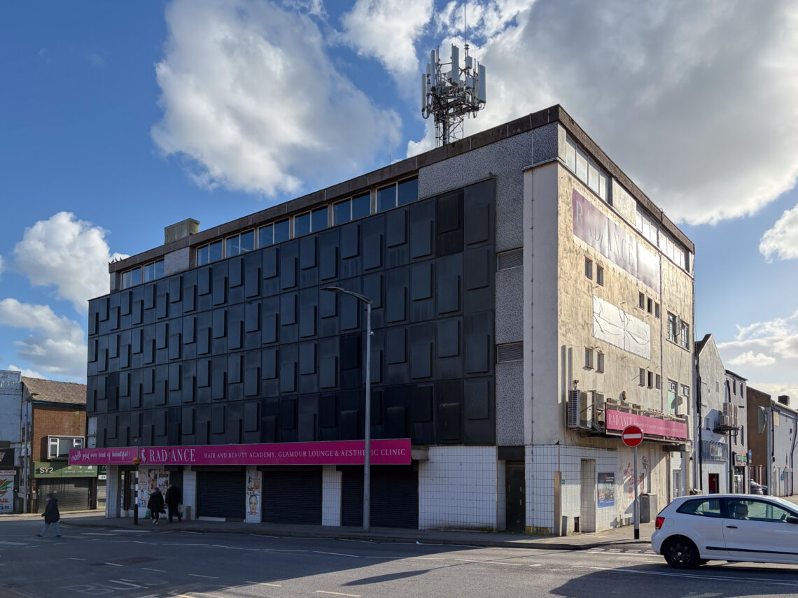 A large, run-down three-storey building with a dark façade, covered windows, and a mobile phone mast on the roof. There is a pink RADIANCE sign for a salon at street level, a few pedestrians passing, and a white car parked nearby. The sky is partly cloudy.