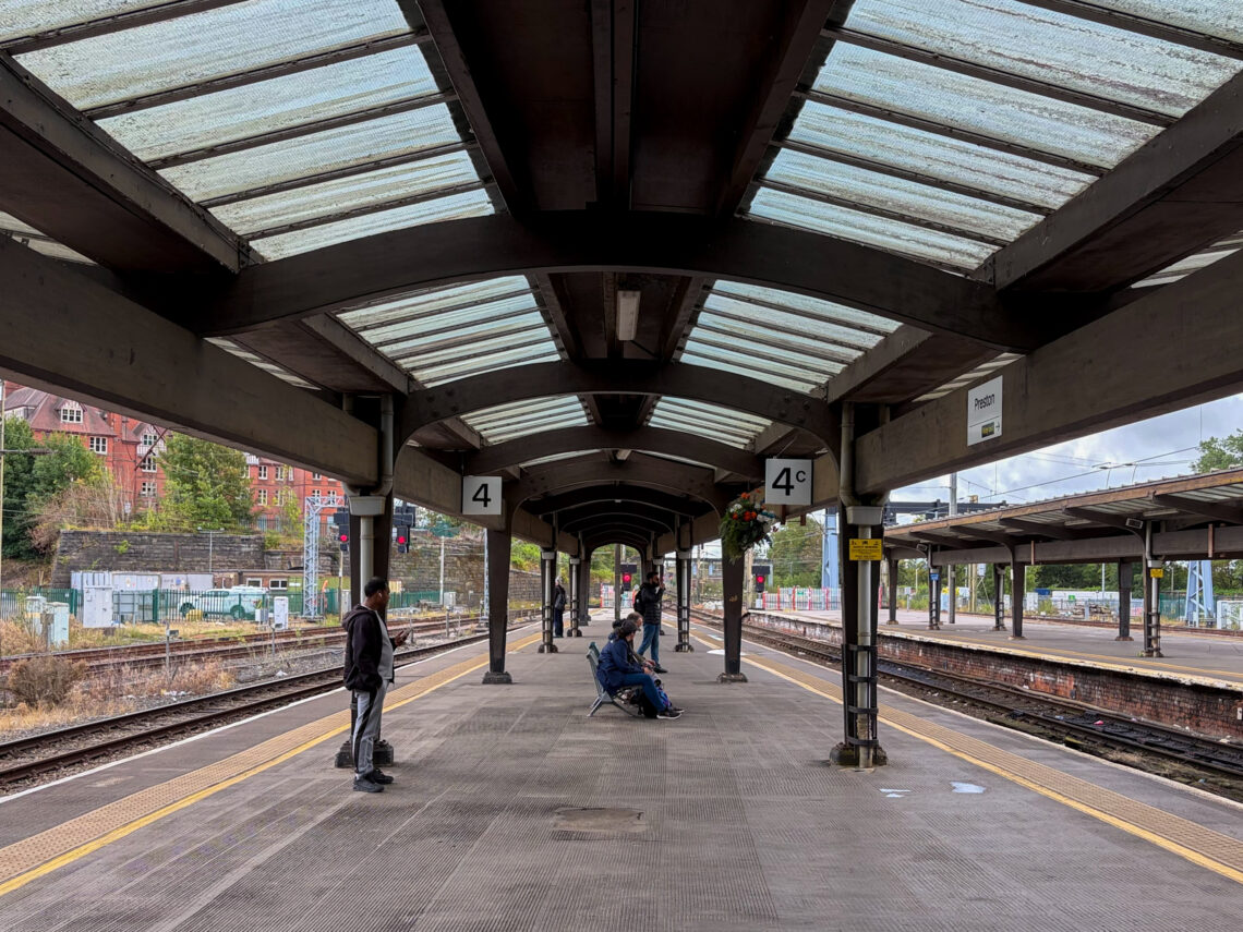 A covered railway station platform with yellow safety lines. Three people wait: one walks, two sit on a bench. Signs show platforms 4 and 4C. Overhead glass panels let in daylight. Tracks and brick buildings are visible in the background. Trees line the area.