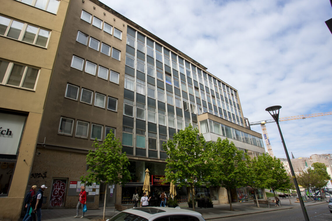 A multi-storey commercial building with a grid of rectangular windows dominates the scene above a ground-floor shopfront. Several young trees line the pavement in front of the building, where a few pedestrians walk near the entrance under a cloudy blue sky.