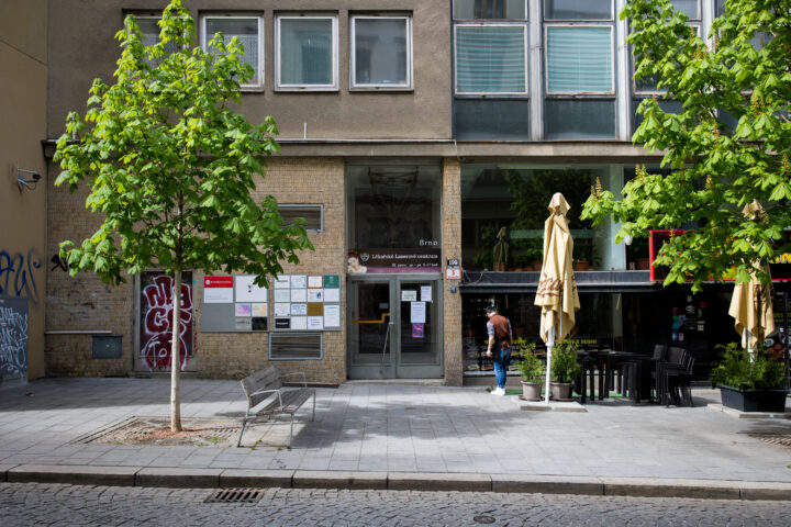An entrance to the Lékářské Laserové centrum in Brno features a glass door with signage and a wall covered in small informational posters. To the right, a person walks past a patio with closed cream-coloured umbrellas and black tables, framed by two leafy trees on a paved pavement.