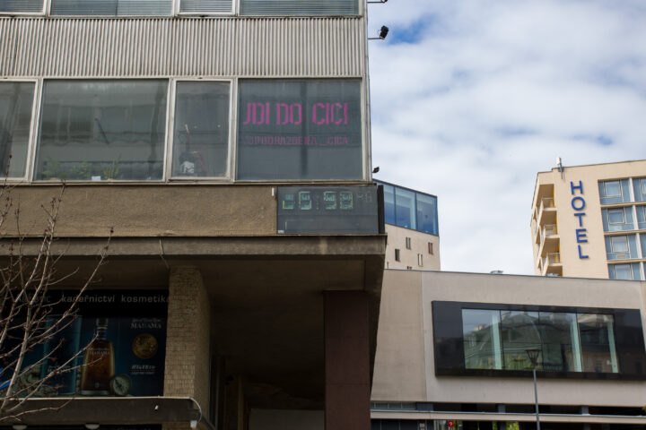 A brutalist building corner features a digital clock displaying 15:58 and a large window with neon pink text reading JDI DO CICI and @PODRAZDENA_CICA. Below, an advertisement for Marama rum is visible on a concrete pillar, with a modern hotel building in the background under a cloudy sky.