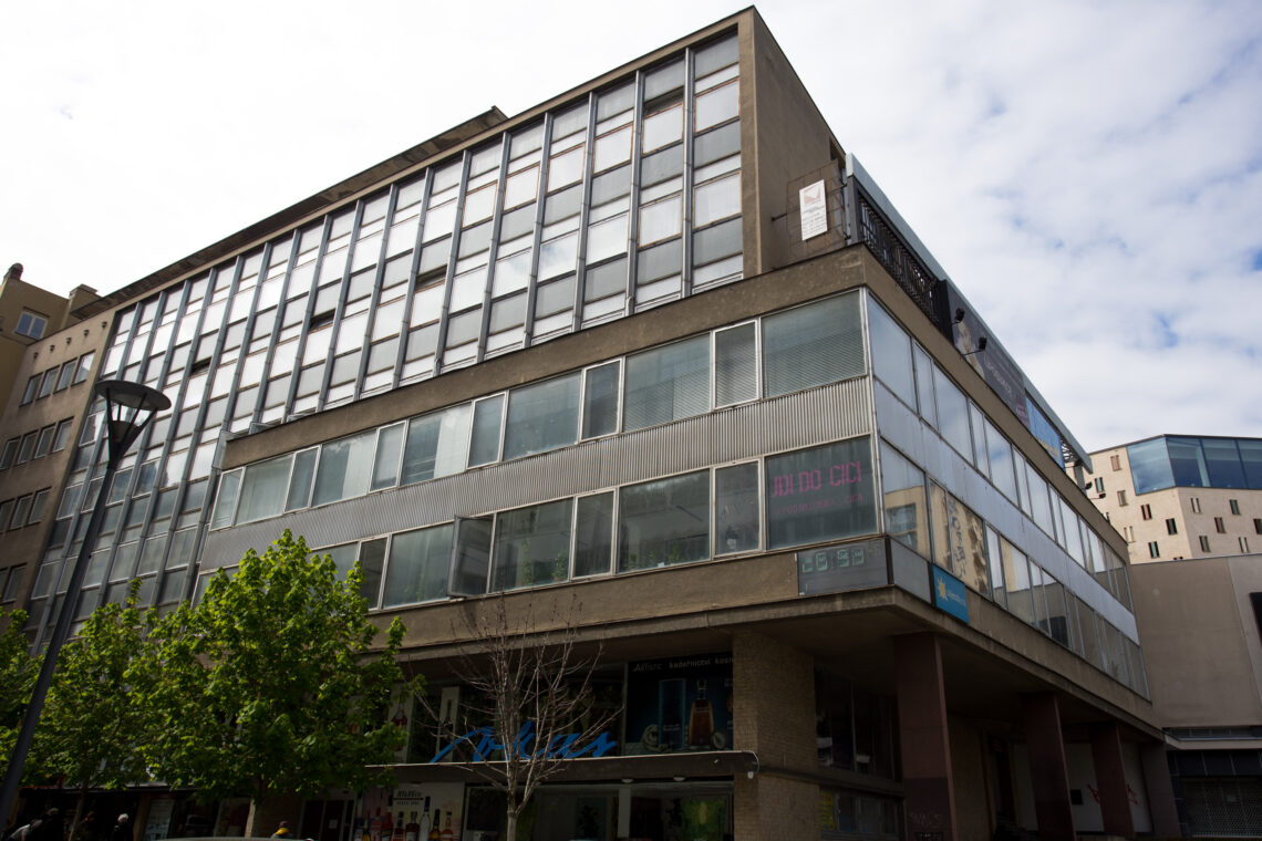 A multi-storey modernist building with a long, glass-panelled façade stands against a cloudy sky. The structure features a grid of windows, a partially obscured neon sign reading IDIO CICI, and street-level shopfronts partially shaded by a leafy green tree.
