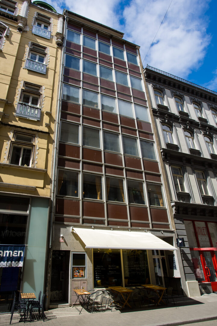A narrow, six-storey modern building with a brown grid façade sits sandwiched between two ornate, historic European-style buildings. At street level, a white awning shades an outdoor café with small wooden tables and chairs set on the pavement. A bright blue sky with wispy clouds is visible above.