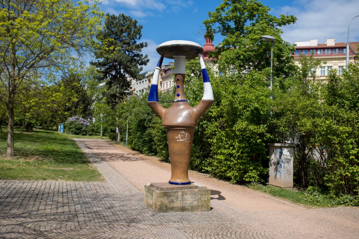 A large, ceramic-style sculpture stands on a stone plinth in a park, featuring a brown vase-like body with blue and white accents supporting a flat, disc-shaped top. It is positioned on a path between green trees and bushes, with historic buildings visible in the background.