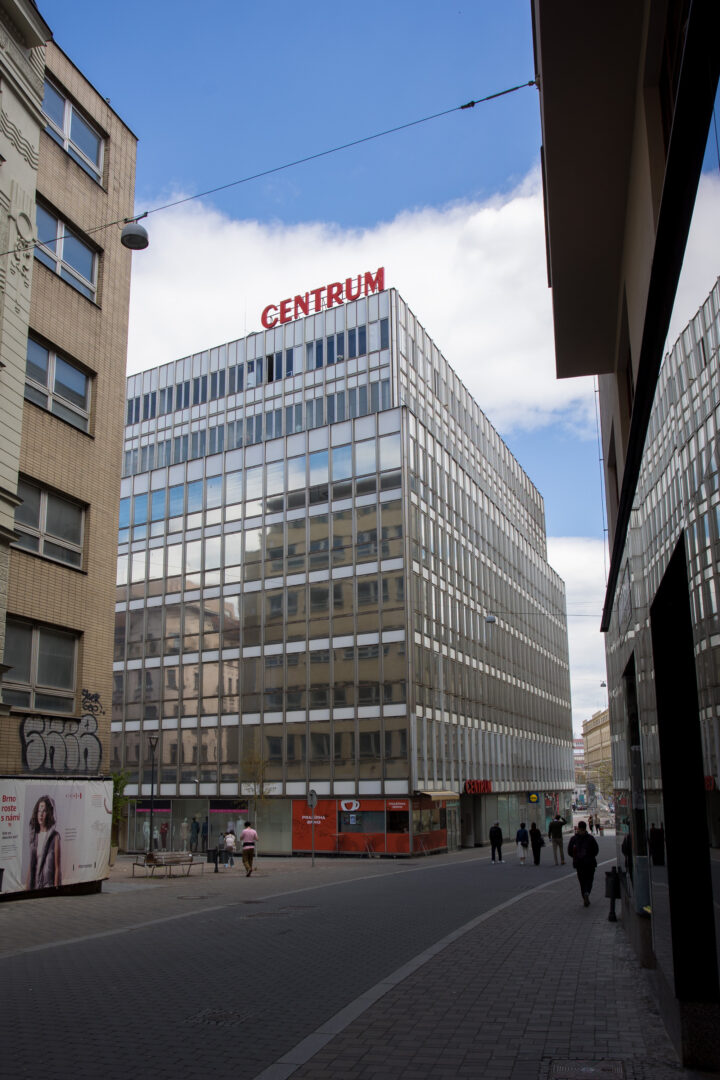 A wide view shows the tall, glass-fronted Centrum building standing in a city square, with CENTRUM in bold red letters on its roof. Pedestrians walk along the paved street in the foreground, flanked by buildings with tan and dark walls under a partly cloudy blue sky.