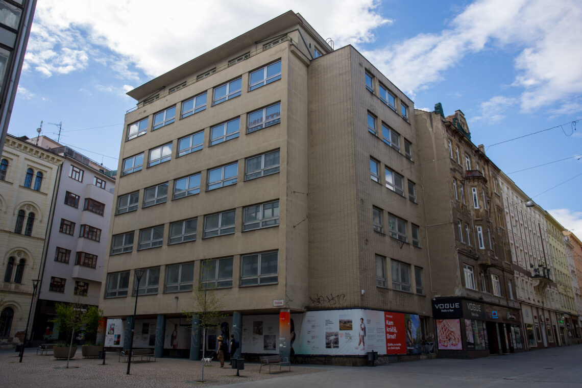 A multi-storey, beige office building with grid-patterned windows sits on a street corner, bordered by older, ornate architecture. At the ground level, white panels cover the base of the building, featuring small advertisements and posters near a few pedestrians walking on the stone plaza.
