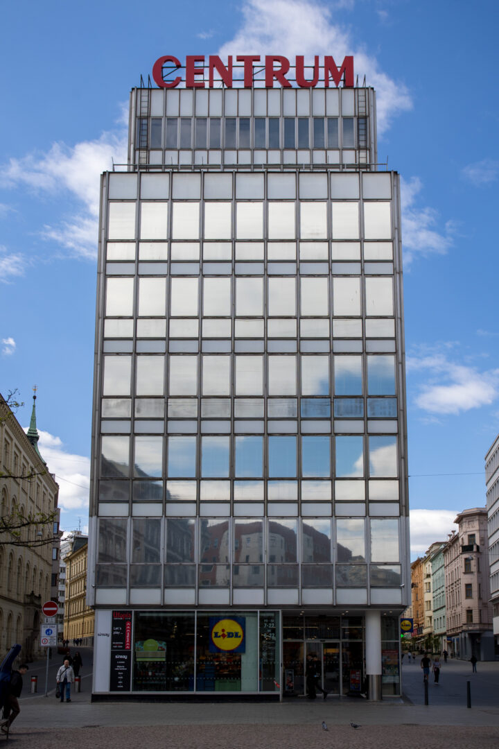 A modern, grid-like glass office building stands under a bright blue sky, topped with a large red sign reading CENTRUM. The ground floor features a Lidl supermarket entrance, whilst the reflective glass facade mirrors the surrounding city buildings and clouds.