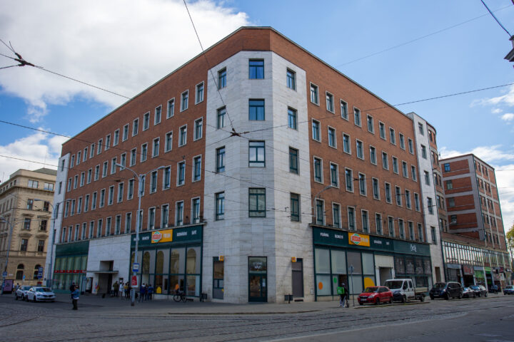 A multi-storey corner building with a red-brick façade and a white stone-clad corner section stands at a city intersection. The ground floor features shopfronts with green frames and Sklizeno signs, while tram tracks curve across the foreground under a cloudy blue sky.