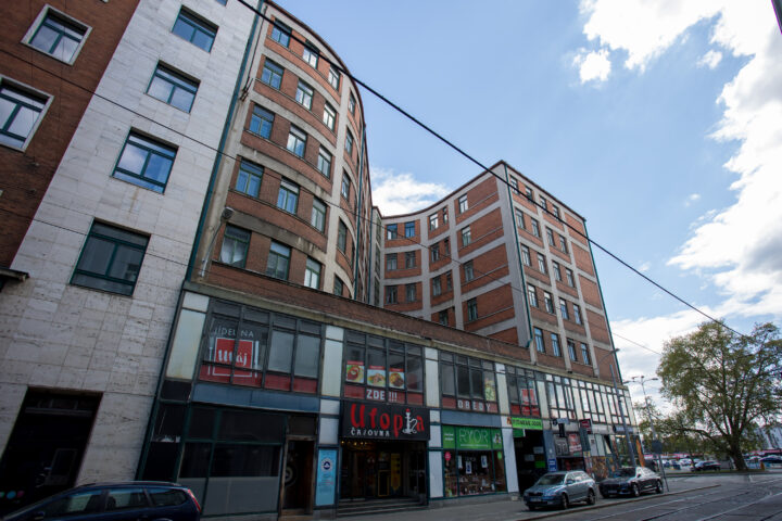 A curved block of flats with a red-brick and white-tiled façade stands against a blue sky with scattered clouds. The street-level shopfronts feature signs for Utopia Čajovna, Ryor, and a Fitness 2000, with cars parked along the pavement in the foreground.