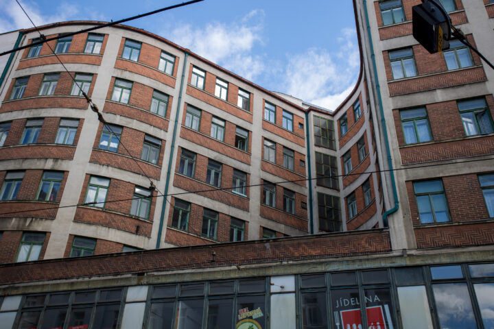 An urban building with a curved façade of red brick and light-coloured concrete stands against a blue, cloudy sky. Multiple rows of windows are visible, and the ground floor features shopfront windows with signs that read Jídelna and Bez lepková. Black trolley wires cross the upper frame.