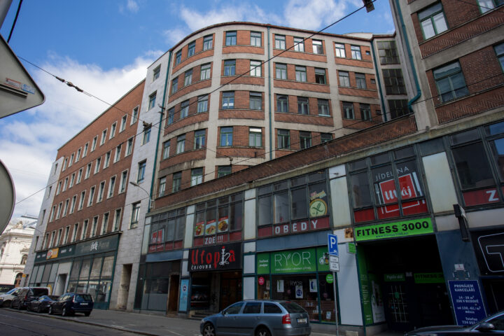 A multi-storey building with a rounded, red-brick corner façade rises above a street-level commercial area. Shops include a tea room labelled Utopia, a Ryor cosmetics store, and a Fitness 3000 gym, with several parked cars lining the pavement under a bright blue sky.