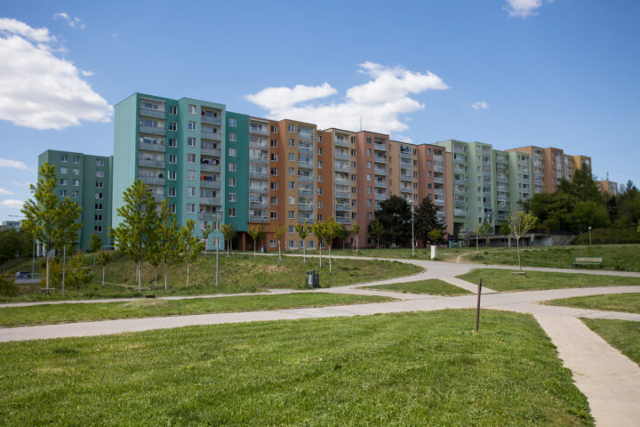 A long, multi-storey block of flats features a colourful facade of alternating turquoise, orange, and light green panels. It sits behind a grassy park with young trees, paved walking paths, and a bright blue sky with scattered clouds above.