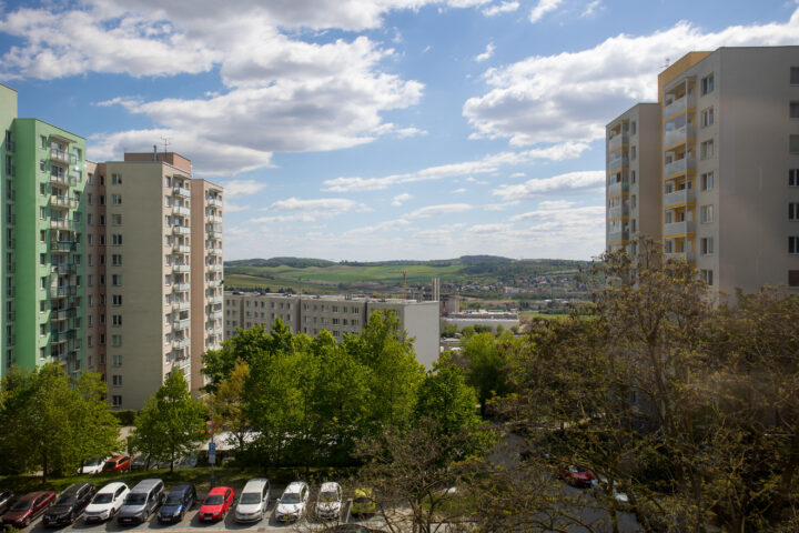 Two tall blocks of flats frame a view of a green valley under a bright, cloudy sky. In the foreground, a car park filled with various cars sits among leafy green trees, while a smaller, long block of flats stands in the distance.